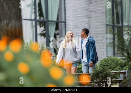 multiethnic couple holding hands and smiling at each other while walking on street on blurred foreground Stock Photo