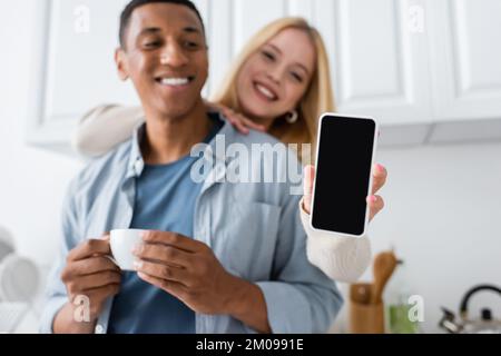 happy blonde woman holding smartphone with blank screen near african american man with coffee cup on blurred background Stock Photo