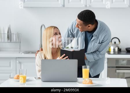 smiling blonde woman showing notebook to african american boyfriend near laptop and breakfast in kitchen Stock Photo