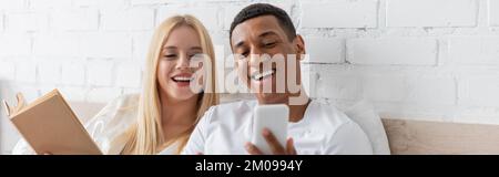 cheerful african american man showing smartphone to smiling girlfriend holding book in bedroom, banner Stock Photo