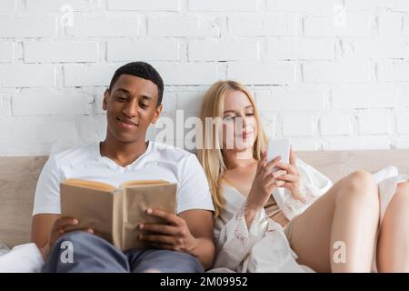 smiling african american man reading book near blonde girlfriend with smartphone in bedroom Stock Photo