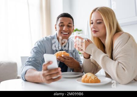 cheerful african american man showing blurred smartphone to smiling girlfriend during breakfast in kitchen Stock Photo