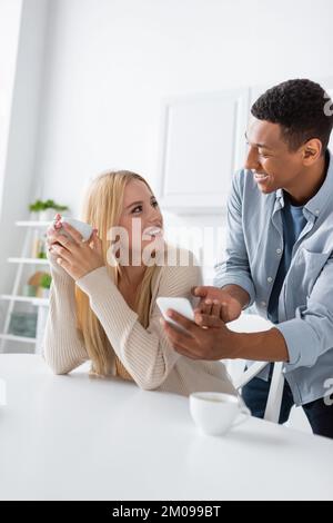 cheerful african american man showing smartphone to blonde woman holding coffee cup Stock Photo