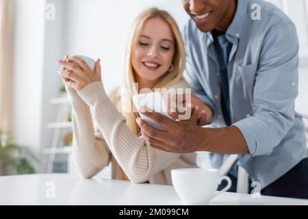 smiling african american man showing cellphone to blurred girlfriend sitting with coffee cup in kitchen Stock Photo