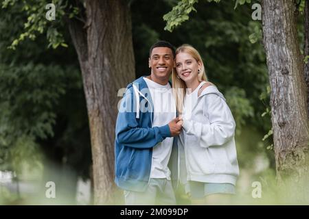 pleased interracial couple holding hands and looking away while standing in park Stock Photo