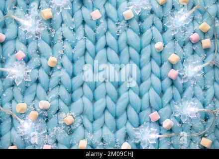 Above view of light blue soft knitted pattern wool background with snowflake shape string lights, fake snow and mini marshmallows, Christmas winter. Stock Photo