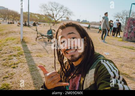young caucasian venezuelan man with dreadlocks making selfie photo with his phone, smiling with good attitude outdoors, walking in the park at sunset, Stock Photo