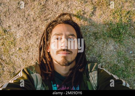 Selfie portrait of young caucasian venezuelan man with dreadlocks and beard, lying on the grass in the park looking at the camera, resting during suns Stock Photo