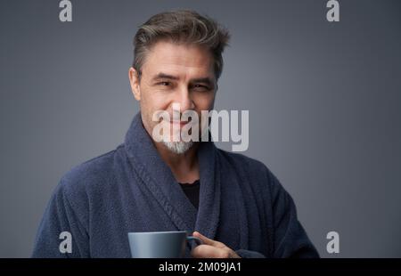 Portrait of mature age, middle age, mid adult casual man in gray bathrobe drinking morning coffee, happy confident smile. Gray background, copy space. Stock Photo