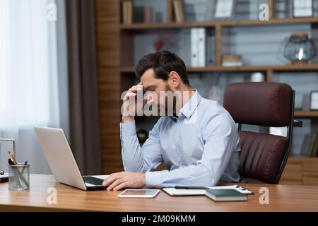 Mature pensive sad businessman working inside office, boss using laptop at work thinking about difficult decision in shirt. Stock Photo