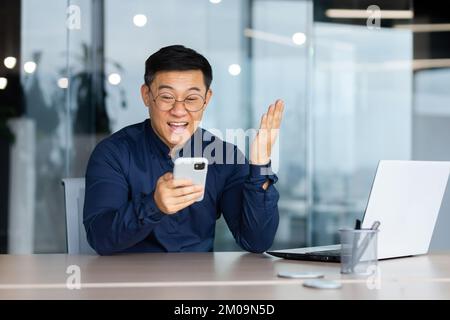 Successful asian businessman celebrating victory and happy good news notification from phone reading, man working inside office using laptop at work holding hand up triumph gesture. Stock Photo