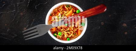 Ramen cup, instant soba noodles in a plastic cup with vegetables, green peas and carrot, overhead flat lay panoramic shot with a fork Stock Photo