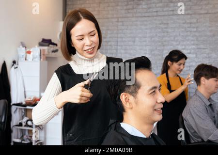 Asian female hairdresser making haircut for two male customer with male hairstyle in qualified barbershop. Men's hairstyling by a scissor and comb in Stock Photo
