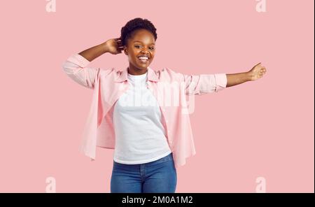 Cheerful young curly laughing afroamerican woman in pink shirt, jeans dancing on pink background. Stock Photo