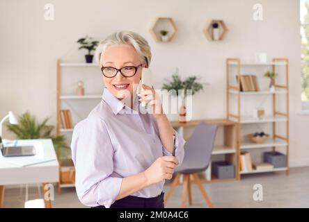 Friendly senior business woman having business conversation on mobile phone in her office. Stock Photo