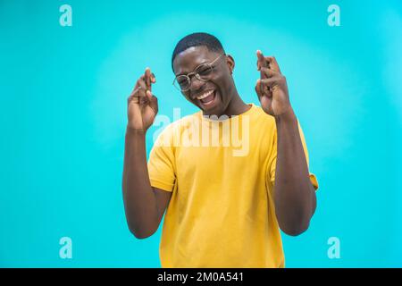 Portrait of a black excited man holding fingers crossed for good luck isolated over blue background. Stock Photo