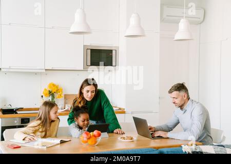 Happy family on cozy kitchen at home, mother helping daughters with homework, father working with laptop Stock Photo