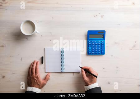 Top view of businessman writing in blank spiral notepad with blue calculator and cup of coffee on the wooden desk. Stock Photo