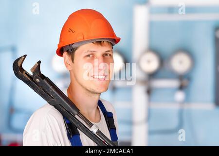 Positive industrial plumber or worker in helmet holds large adjustable wrench on his shoulder and smiles. Indoor portrait against background of sensors and pressure gauges in blur.. Stock Photo