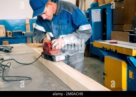 Professional woodworker grinds wooden bar on workbench in carpentry workshop with tool. Middle-aged worker in overalls processes wood with electric grinder. Real workflow.. Stock Photo