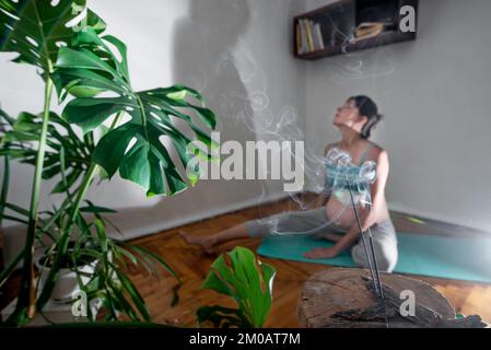 Pregnant woman sits on a yoga mat in a meditation pose. Pregnant woman doing yoga exercises at home. Incense room for relaxation during yoga Stock Photo