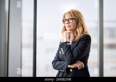Mature female looking deep in thought while standing with arms crossed. Confident businesswoman wearing eyewear and blazer. Copy space. Stock Photo