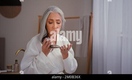 Caucasian old sad senior mature woman lady grandmother model in bathrobe in bath touching gray long smooth thin hair worry about lost hairline weak Stock Photo