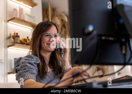 Positive adult female employee with long dark hair in casual wear and eyeglasses smiling and typing on computer keyboard while having phone call  Stock Photo