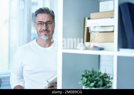 Portrait of mature age, middle age, mid adult casual man in glasses at home, happy smiling, gray hair, copy space. Standing at book self reading. Stock Photo