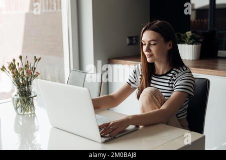 Pensive, smiling young brown long haired woman sitting in white kitchen, use laptop. Freelancer and self-employed worker Stock Photo