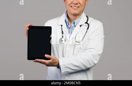 Smiling adult japanese guy therapist in white coat with stethoscope shows tablet with empty screen Stock Photo
