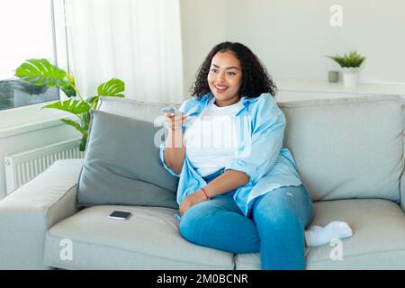 Happy african american plus size woman watching TV, switching channels with remote controller, sitting on sofa at home Stock Photo