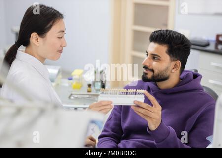 Tooth teeth tone palette. Beautiful asian female stomatologist orthodontist dentist choosing selecting color of patient attractive bearded man teeth for whitening at clinic. Stock Photo