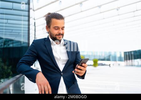 Businessman standing outdoor use smartphone looking device screen and smile. Handsome hispanic male business person middle age dressed business suit holding smartphone in hand. Modern business person Stock Photo