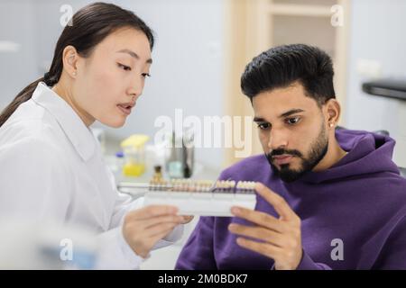 Tooth teeth tone palette. Beautiful asian female stomatologist orthodontist dentist choosing selecting color of patient attractive bearded man teeth for whitening at clinic. Stock Photo