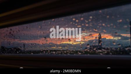 Looking out at sunrise from the inside of a campervan on a rainy day through the blinds towards Flamborough Head lighthouse and beacon on the headland Stock Photo