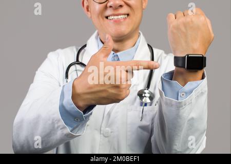 Happy adult asian man doctor in white coat pointing finger at watch with empty screen Stock Photo