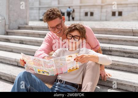 Tourist Couple With Map In Capital City Europe. Happy Young People Using Map, Traveling On Summer Vacations. Handsome Man And Beautiful Woman Having Trip Outdoors. High Quality Image. Sitting stairs Stock Photo