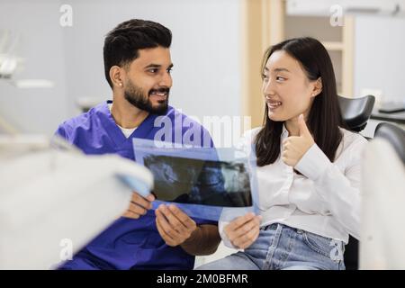 Medicine, dentistry and oral care concept. Male confident dentist showing x ray picture to attractive young patient, asian woman at modern light dental clinic. Stock Photo