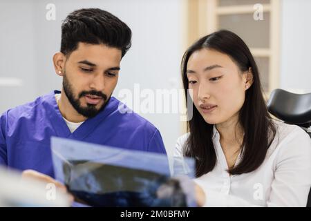 Medicine, dentistry and oral care concept. Male confident dentist showing x ray picture to attractive young patient, asian woman at modern light dental clinic. Stock Photo