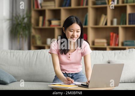 Cheerful korean lady attending webinar from home, sitting on sofa, using laptop and taking notes, copy space Stock Photo