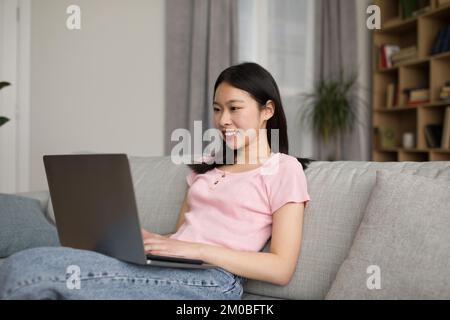 Happy korean woman freelancer sitting on sofa, typing on laptop keyboard and smiling, home interior, free copy space Stock Photo