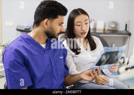 Attractive young asian woman, sitting on dentist chair and looking at x-ray picture of scan image of teeth together with her confident professional male dentist at clinic. Stock Photo