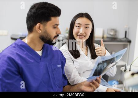 Attractive young asian woman, sitting on dentist chair and looking at x-ray picture of scan image of teeth together with her confident professional male dentist at clinic showing thum up. Stock Photo