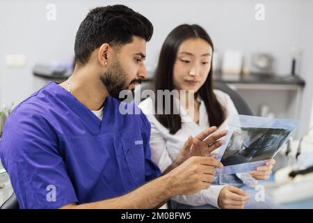 Attractive young asian woman, sitting on dentist chair and looking at x-ray picture of scan image of teeth together with her confident professional male dentist at clinic. Stock Photo