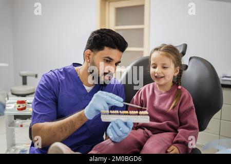 Visual method of subjective perception color of filling by a doctor. Bearded confident male dentist checking and selecting color of young caucasian girl's teeth. Stock Photo