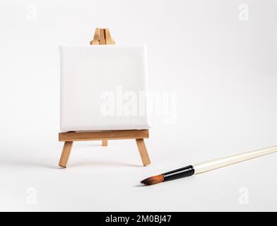 Square canvas on tiny mini easel, blank mockup on wood tripod, paintbrush. Artistic supplies composition. High quality photo Stock Photo