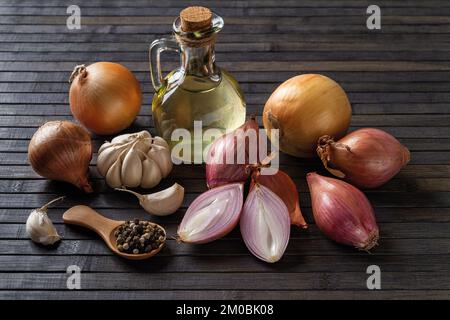 Whole and halved red shallot, yellow onion, garlic, peppercorns and olive oil on a black wooden background. Raw onion bulb for spice and seasonings. Stock Photo
