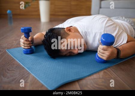 Tired asian mature man lying on fitness mat with dumbbells in hands, resting after domestic workout in living room Stock Photo