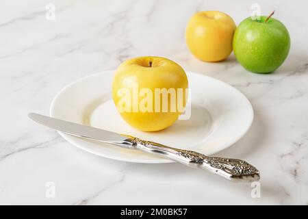 Ripe yellow apple and knife on a white plate over marble background. Fresh juicy fruit for vitamin healthy eating. Antioxidant vegetarian snack. Stock Photo